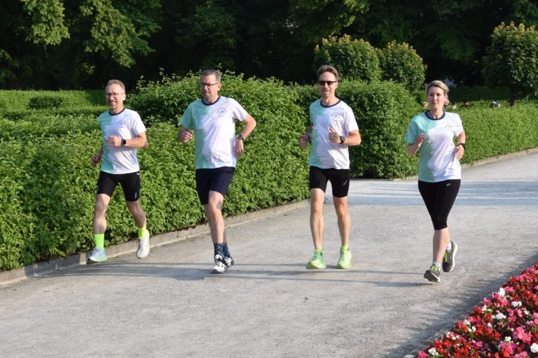 Vier Personen joggen auf einem Weg neben bunten Blumenbeeten in einem Park.