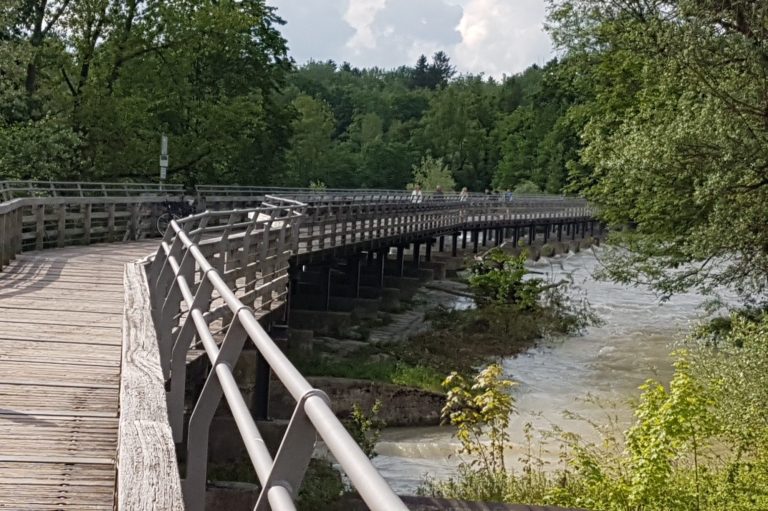 Holzsteg über einen Fluss, umgeben von grünen Bäumen und wolkenlosem Himmel.