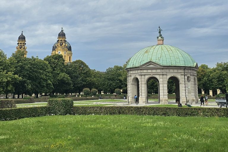 Kleinod im Park mit einem Pavillon und historischen Türmen im Hintergrund.