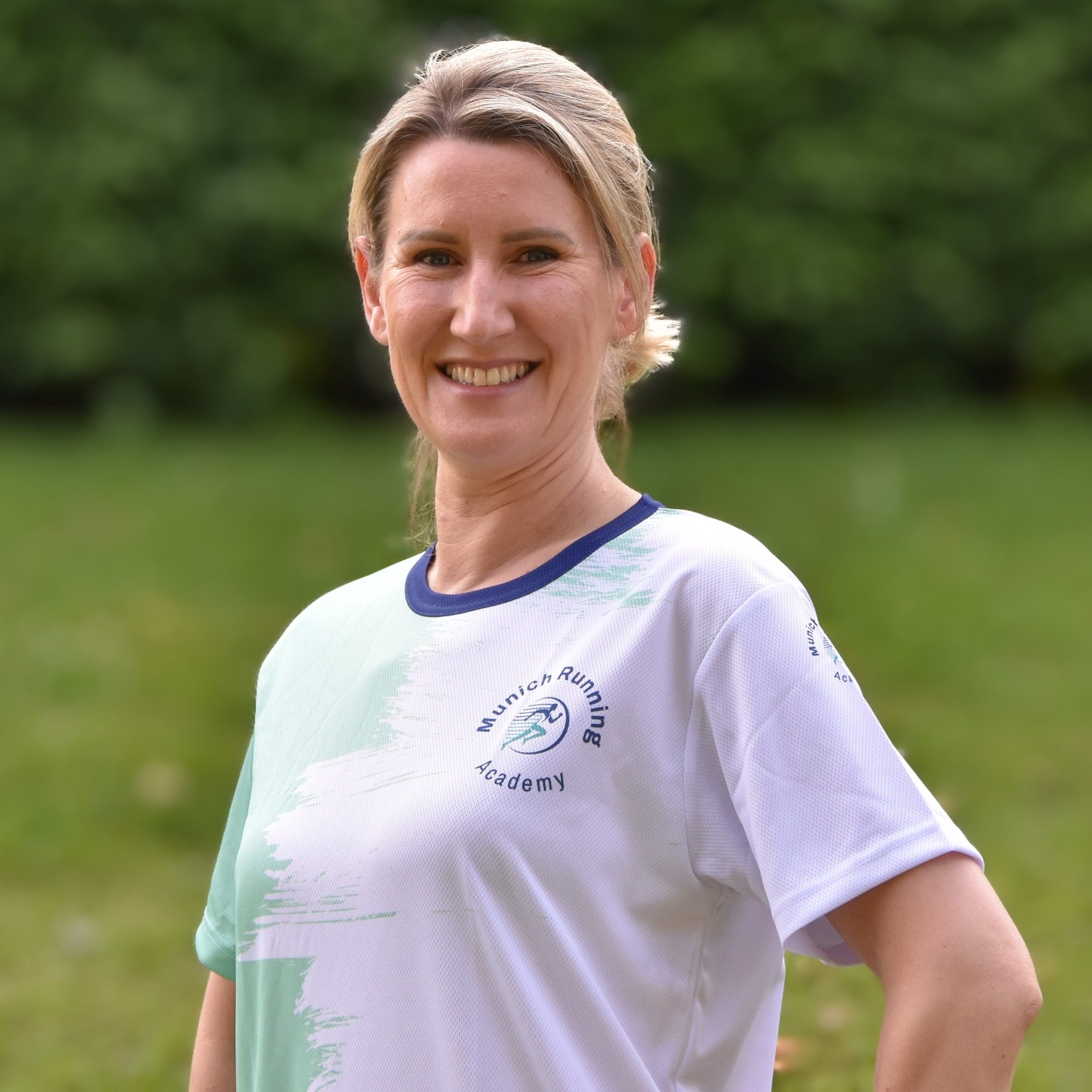 Lächelnde Frau in Sportkleidung, steht in einer grünen Umgebung.