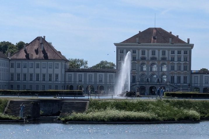 Schloss mit Brunnen und umliegendem Wasser, umgeben von Bäumen und blauem Himmel.