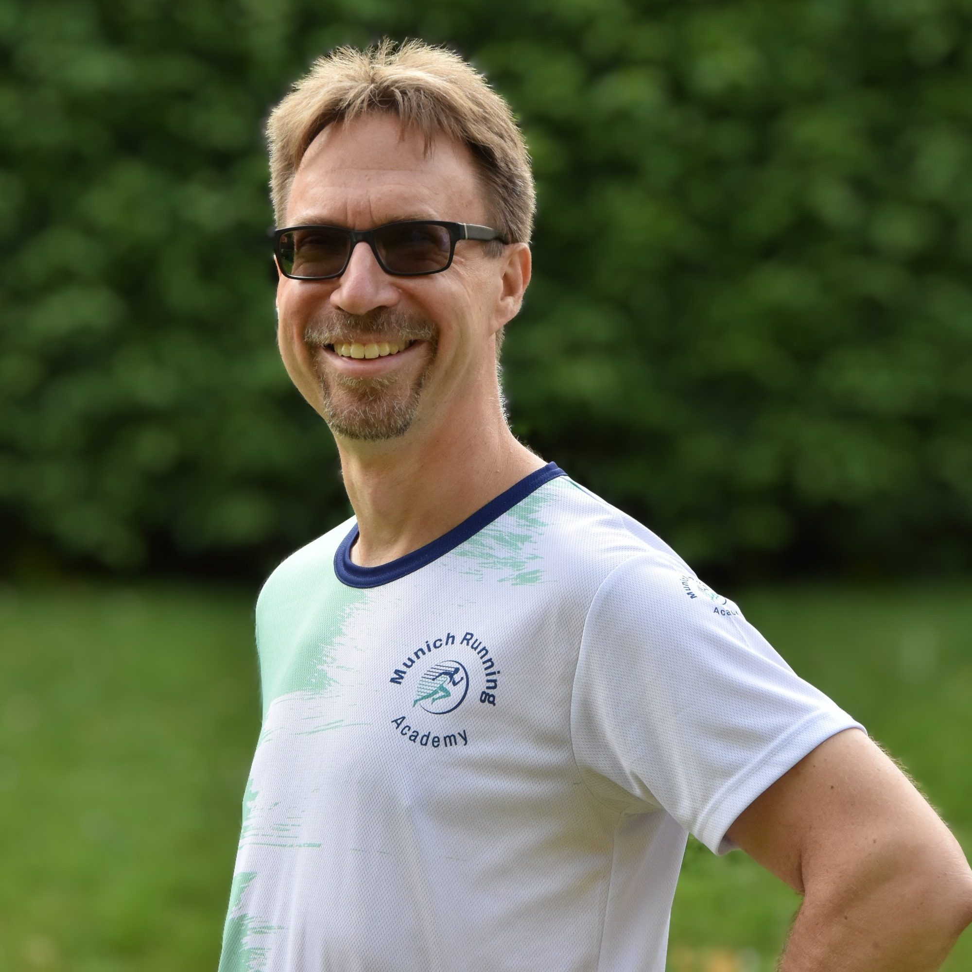 Mann mit Sonnenbrille und T-Shirt steht lächelnd im Freien vor grünem Hintergrund.