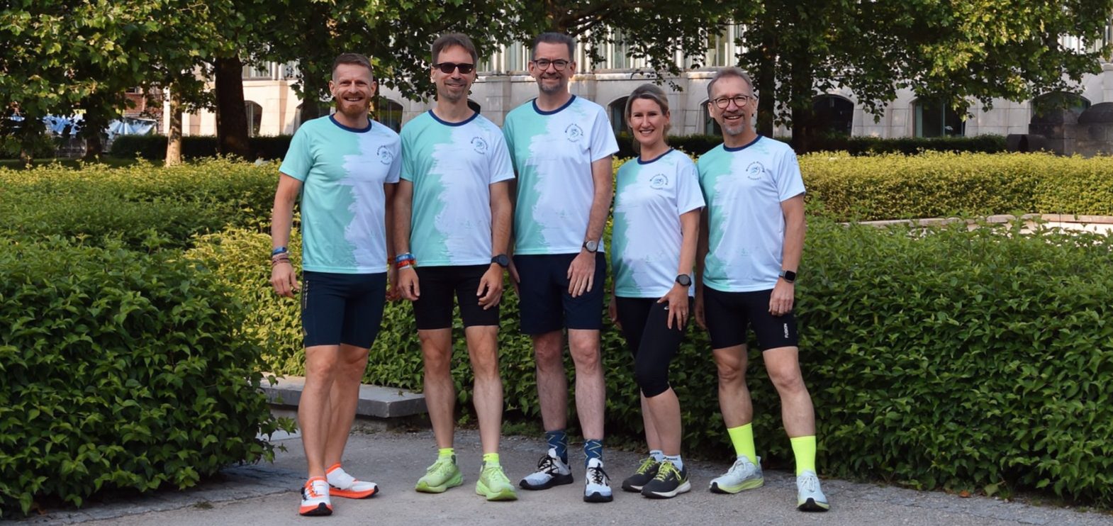 Fünf Personen in Sportkleidung stehen lächelnd vor einer grünen Hecke.