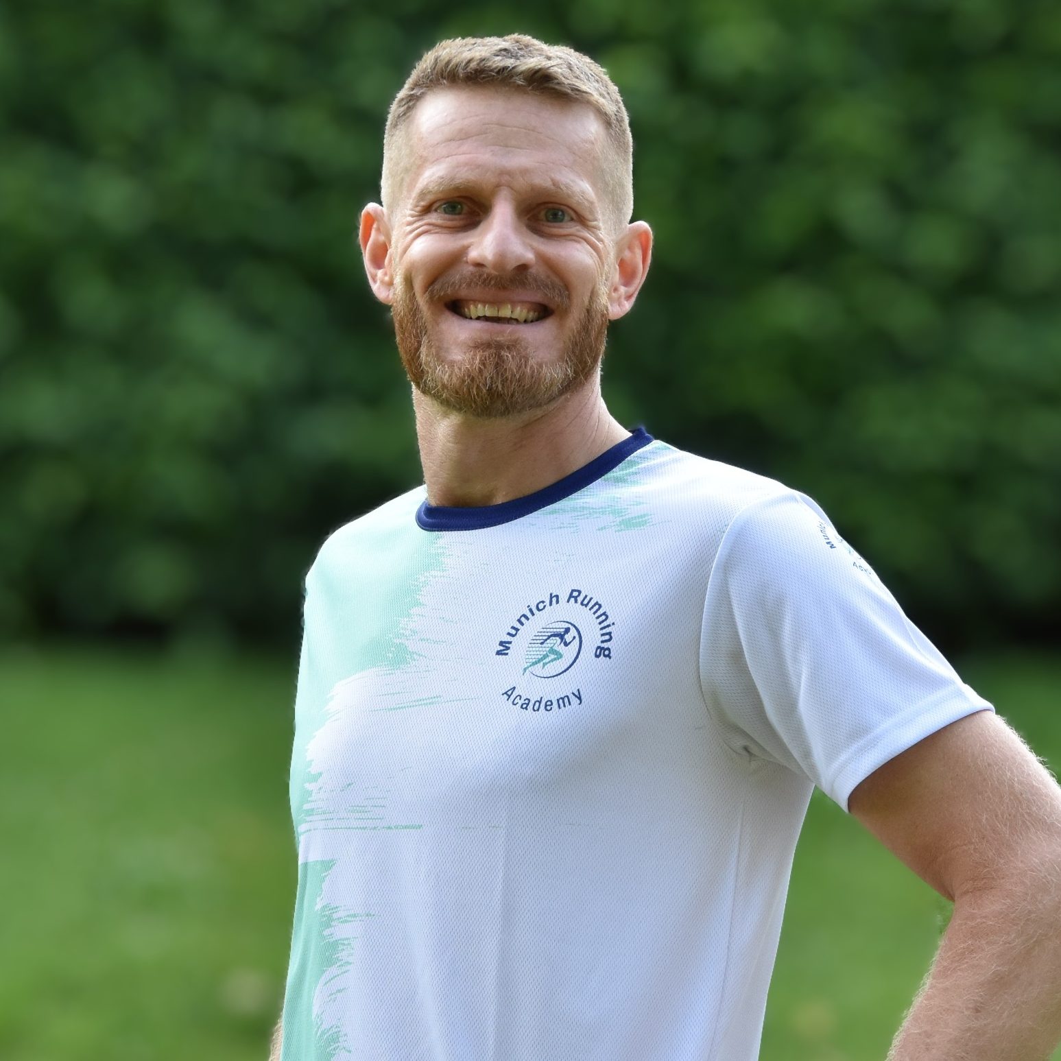 Lächelnder Mann in einem weißen T-Shirt, steht vor einer grünen Kulisse.