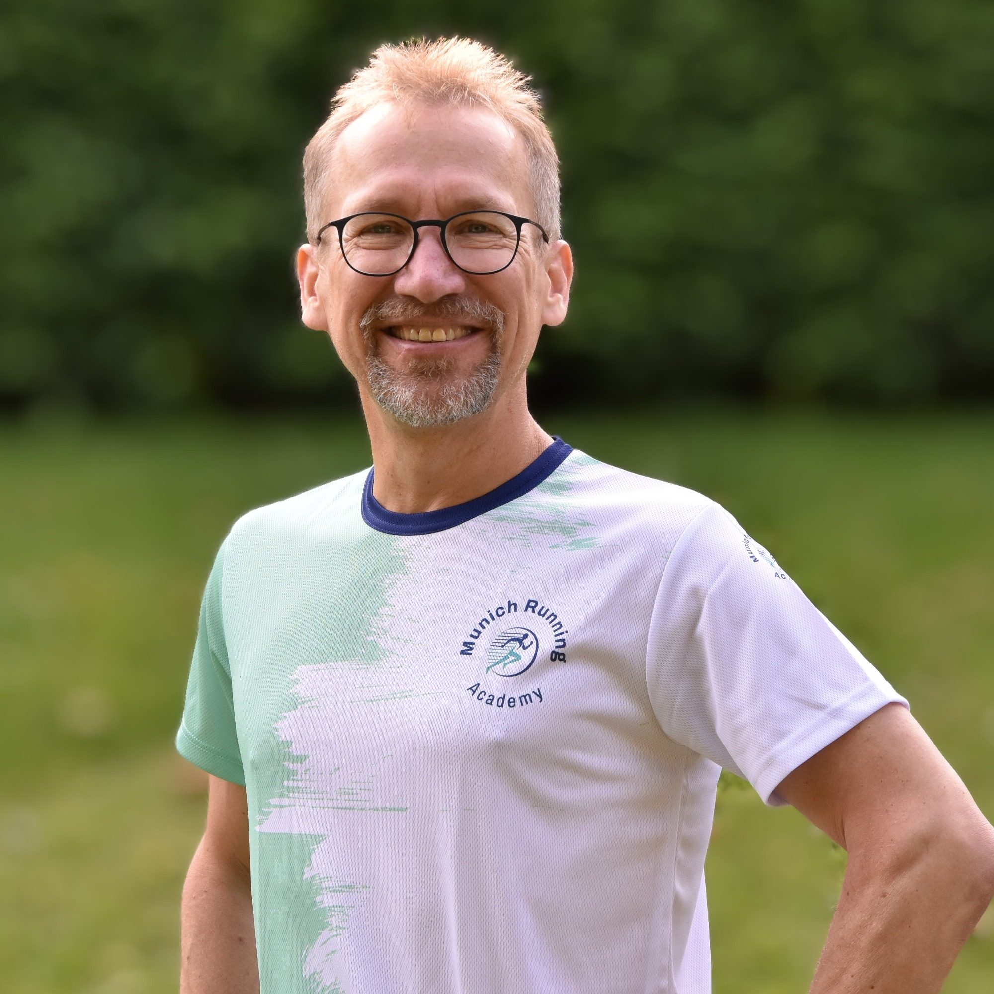 Mann mit Brille, lächelnd, trägt ein T-Shirt in grün und weiß, steht im Freien.